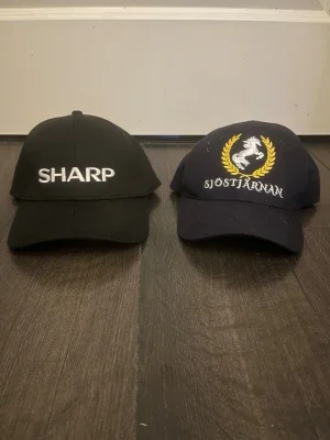 Svart keps med SHARP-brodyr och blå keps med ”sjöstjärnan brodyr”